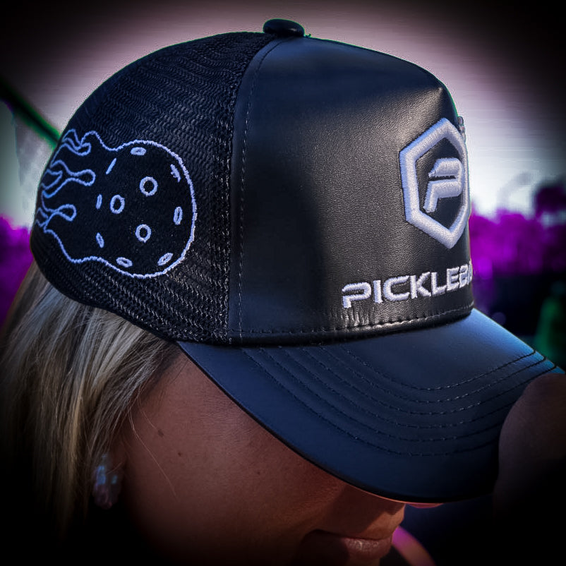 A person wears the Potenza Pickleball Leather Logo Trucker Hat, a black ventilated cap with “PICKLEBALL” and hexagonal logo on the front, and a white embroidered pickleball design on the side. Blonde hair peeks out from underneath.