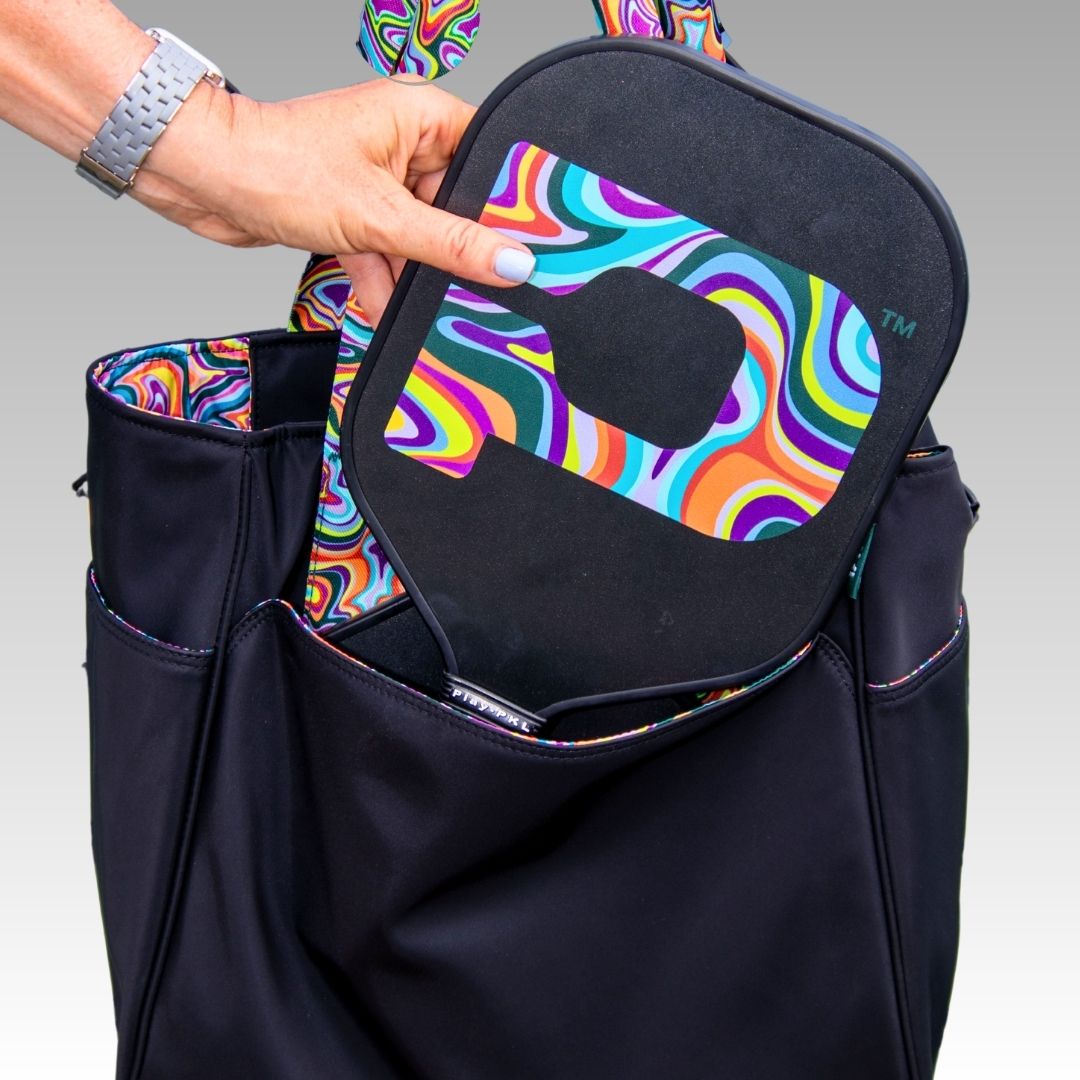 A hand places the Playly Store Signature Pickleball Paddle, featuring a psychedelic P design and graphite surface, into a black sports bag with rainbow stitching and side pockets. The person wears a light gray watch.