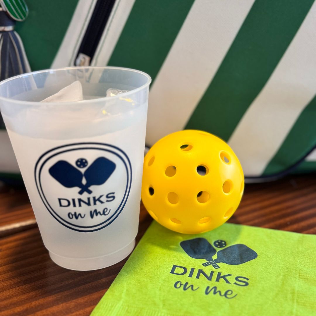 A Fresh Pickle Pickleball Roadie Cup with ice water and a “DINKS on Me” logo sits next to a yellow pickleball, green napkin, and a green-and-white striped bag on a wooden table.