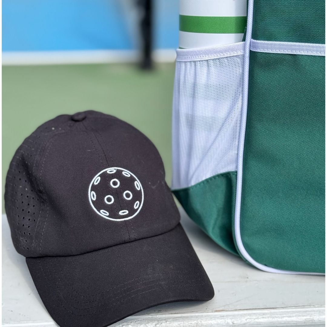 The Fresh Pickle Pickleball Caps, featuring a black design with a white graphic, rest beside a green and white sports bag—perfect as a stylish, breathable cap for women on or off the court.
