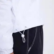 A close-up shows a person in the Runway Athletics Court Jacket, a cozy white fleece with an adjustable drawstring hem, paired with black pants with bold white stitching. The background is plain and light-colored.