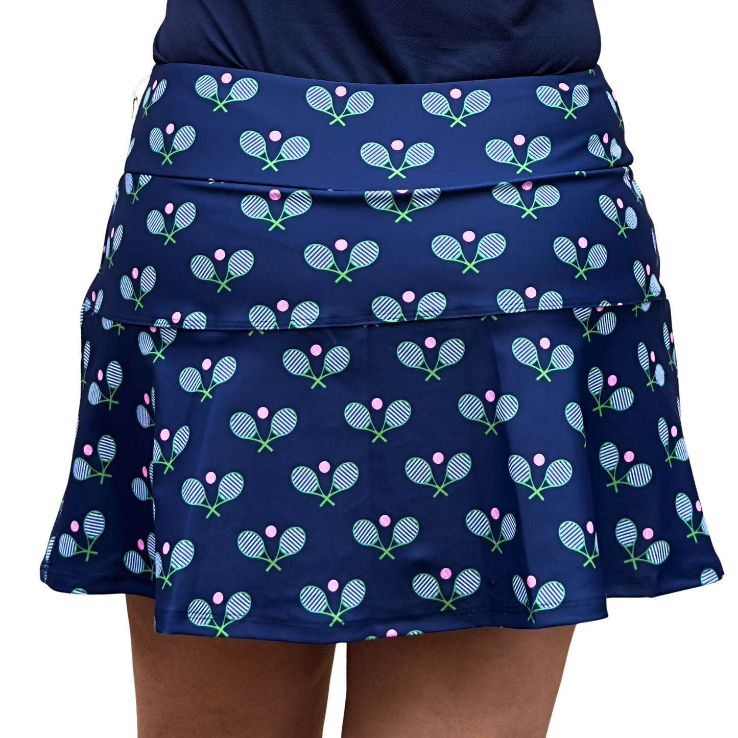 A woman models the Queen of the Court Stripey Tennis Navy skirt, made in the USA, with a ruffled layered design and navy blue color featuring crossed rackets and pink tennis balls. Only her lower torso is shown, paired with a dark top.