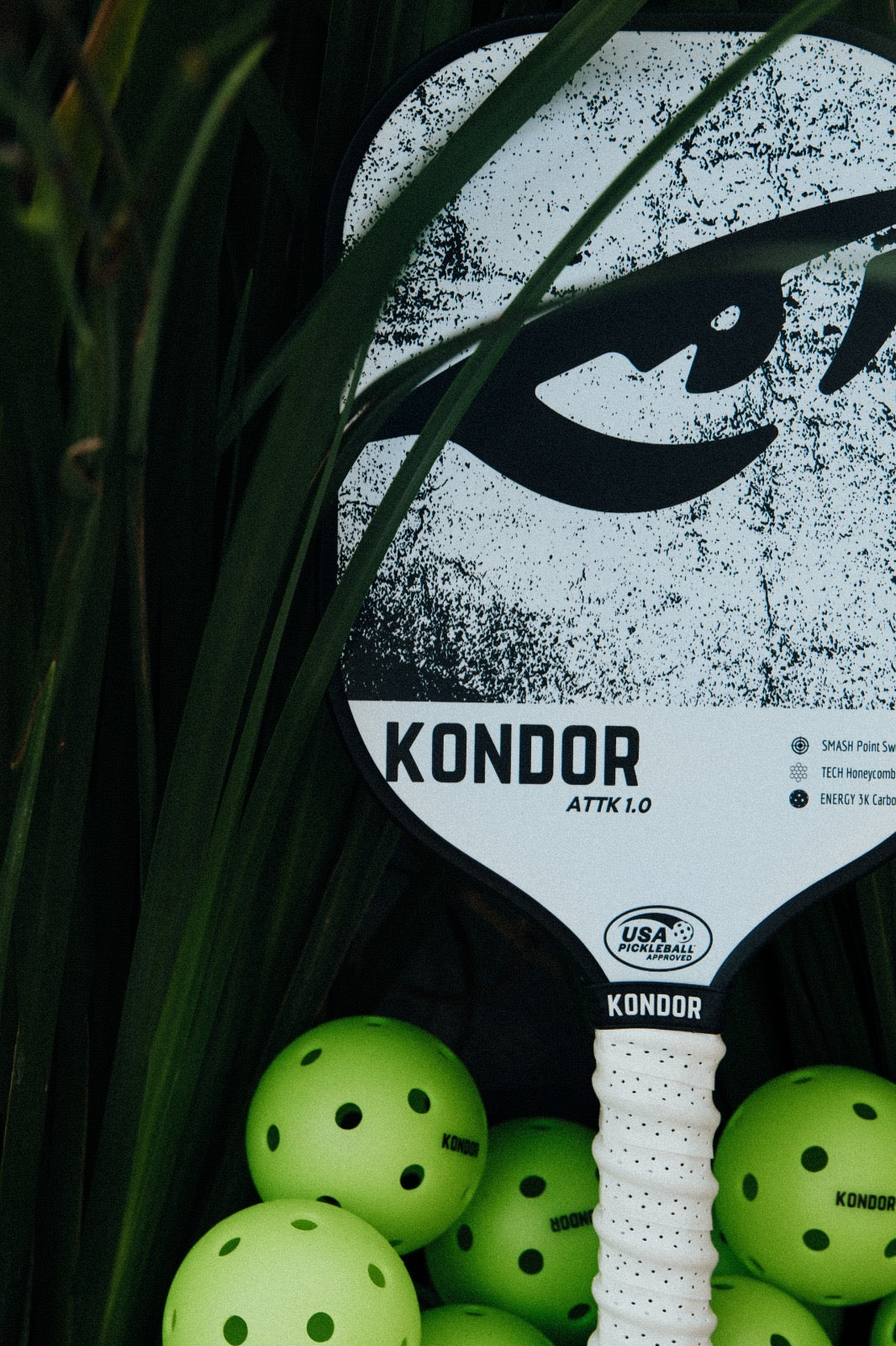 A KONDOR PICKLEBALL ATTK 1.0 paddle with an elongated grip stands among grass-like leaves, with several green perforated pickleballs at its base and the USA Pickleball logo displayed on the paddle.