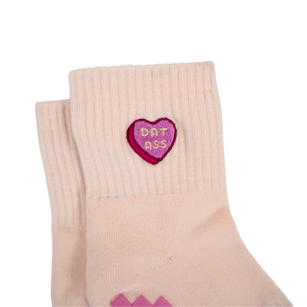 The Sweet Nothings Grip Sock by Revel is a light pink Pilates sock with a ribbed cuff and a small pink-and-red embroidered heart patch featuring "DAT ASS" in yellow letters. Shown laid flat on a white background.