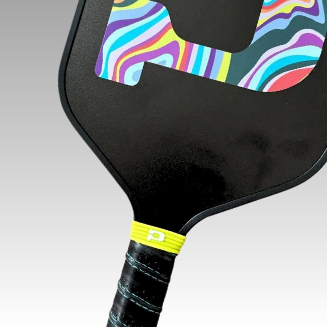 Close-up of a black Playly Store pickleball paddle featuring a colorful wavy "P," with a textured grip wrapped in black and highlighted by the Pickleball Paddle Icon Grip Band—yellow with a white logo—set against a light gray gradient background.