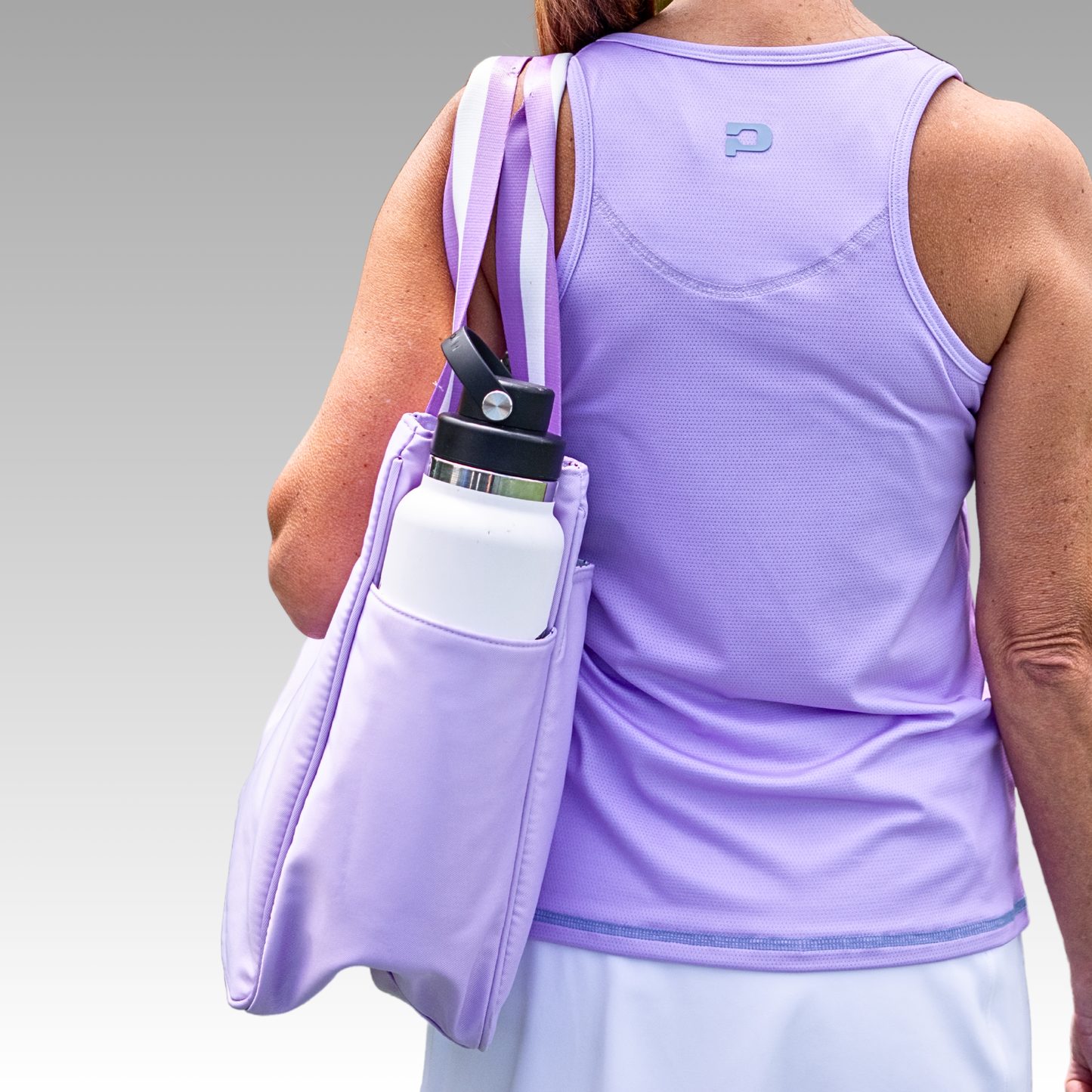 A woman seen from behind wears a sleeveless lavender top, white skirt, and carries the Playly Store’s Lulu Pickleball Tote in matching lavender with a large white water bottle in its side pocket. The background is a neutral gray gradient.