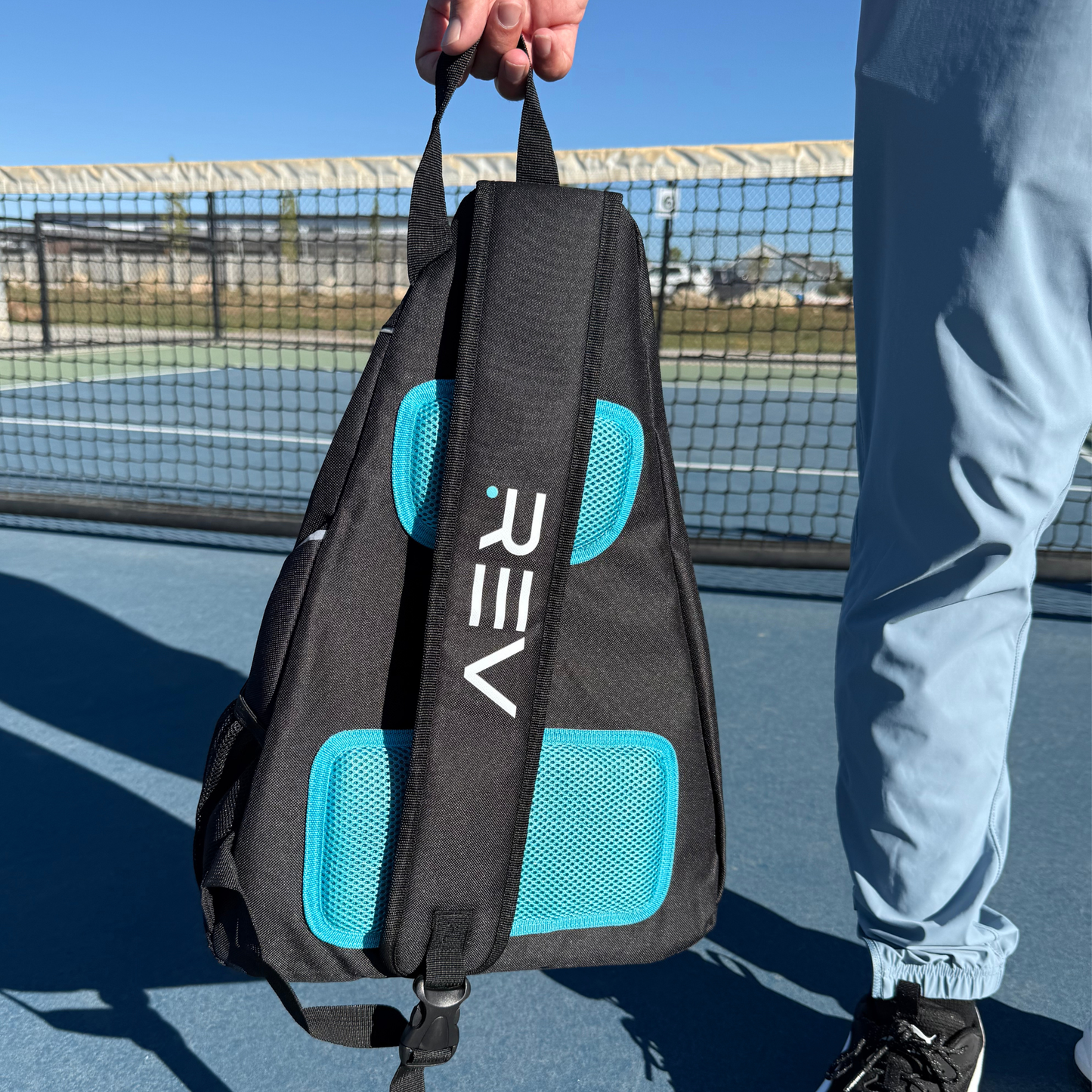 A person in light blue pants holds the REV Pickleball ELITE Paddle Bag, featuring black material and turquoise mesh pockets. "REV" is printed on the strap. They stand outdoors by a tennis court and net on a sunny day.