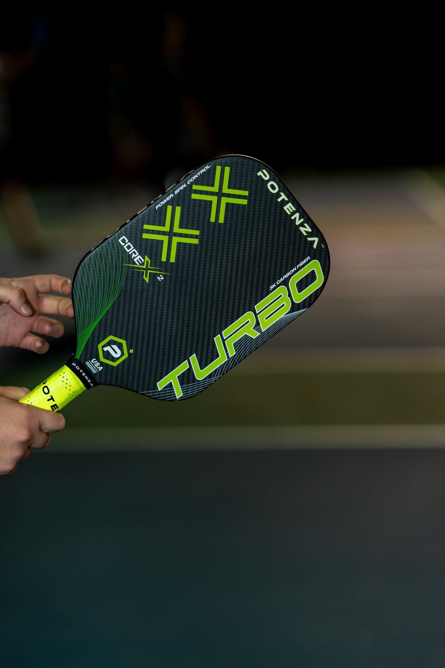A person holds a black and neon green Potenza Pickleball TURBO+ COREx2 paddle with a textured carbon fiber design, standing out over a blurred indoor court. Only the hand and the power paddle are clearly visible in the image.
