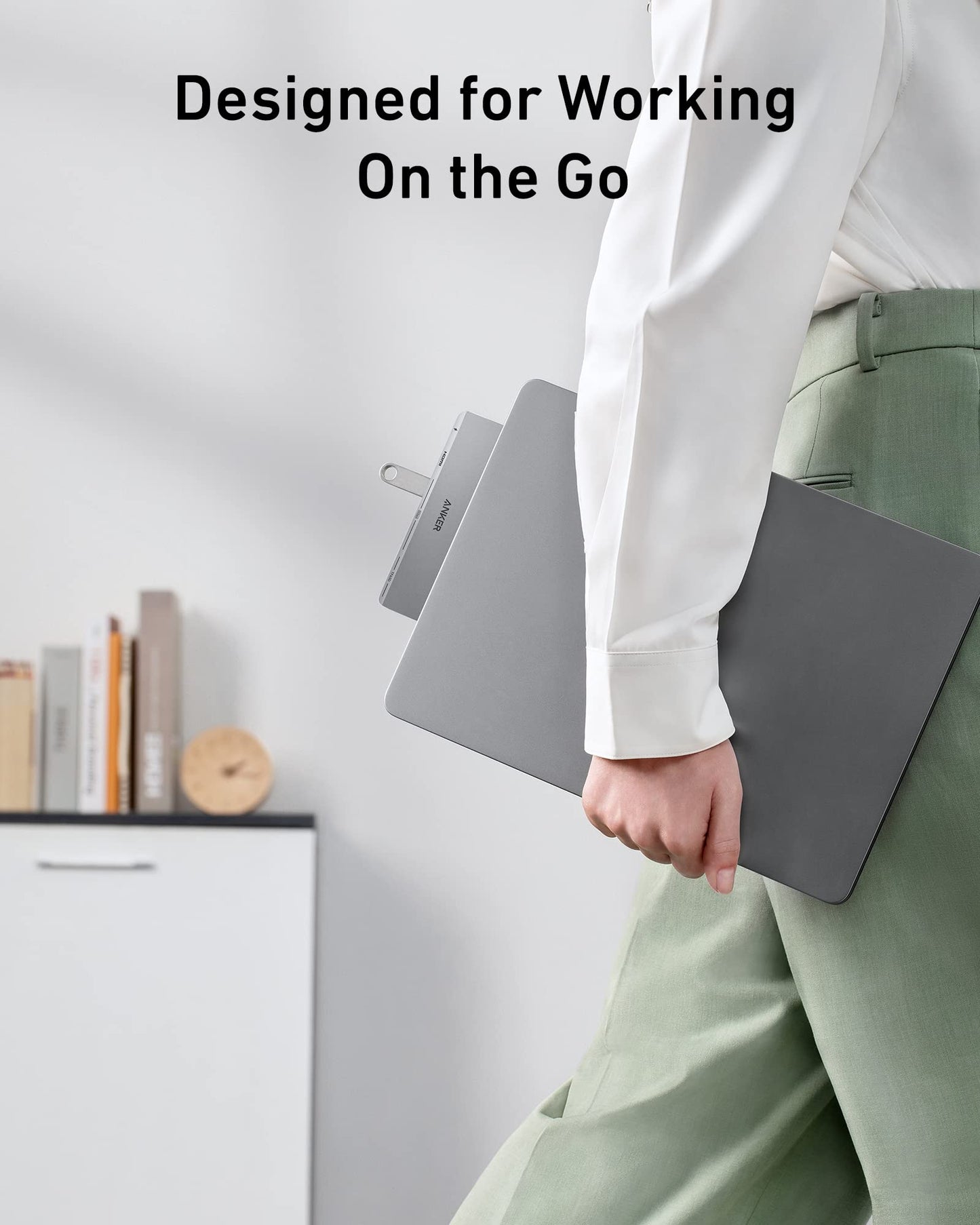 A person in green pants and a white shirt holds a closed gray MacBook with the Anker <b>547</b> USB-C Hub (7-in-2, for MacBook) attached and a USB drive inserted. Behind them is a white cabinet with books and a wooden clock. The text reads: Designed for Working On the Go.