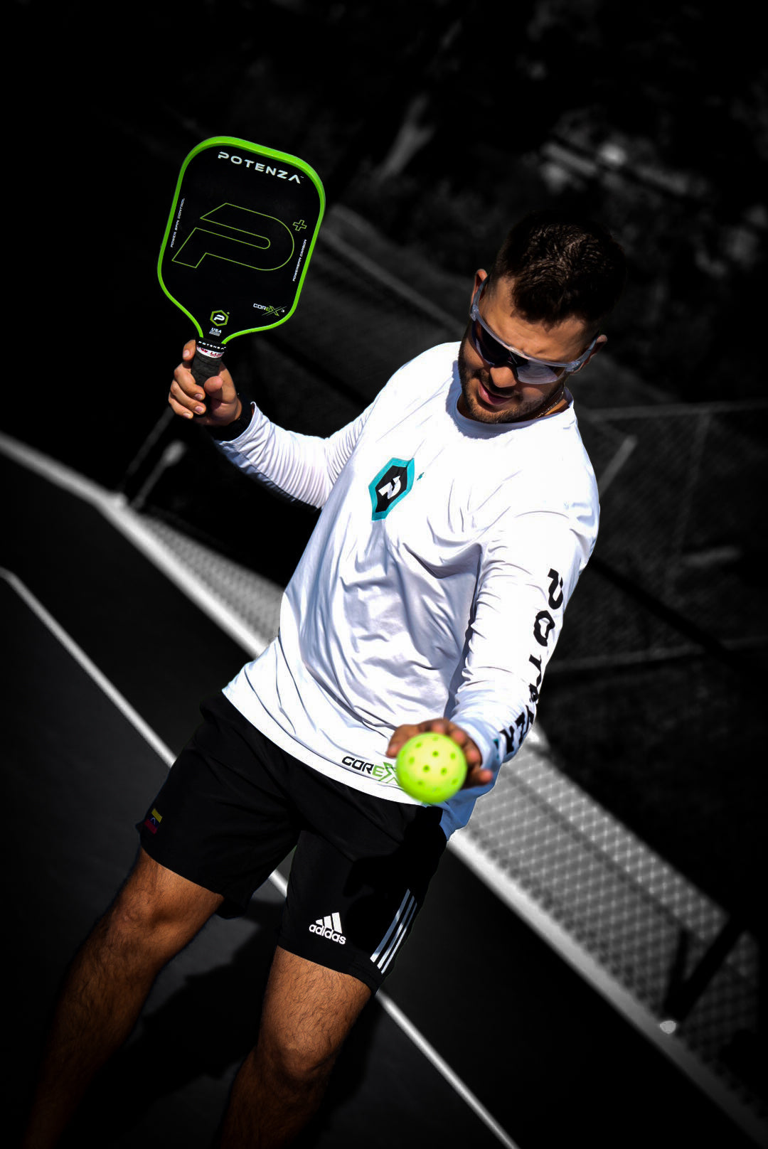 A man in sunglasses, a white long-sleeve shirt, and black shorts stands on an outdoor court holding a Potenza Pickleball P+ PowerSpin Carbon COREx3 (Neon Green) paddle and a yellow pickleball against a black-and-white background.