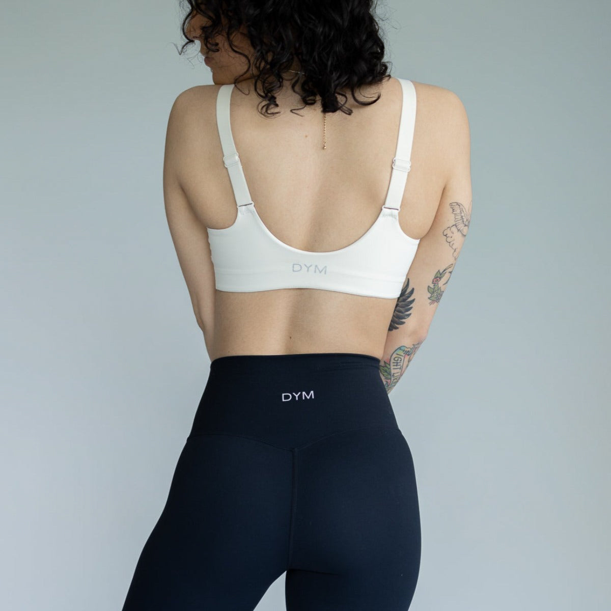 A person with curly dark hair, visible tattoos on their left arm, faces away wearing the white DYM Athletics Element Essential Bra (A-D cup, medium support) and black high-waisted leggings against a plain light grey background.