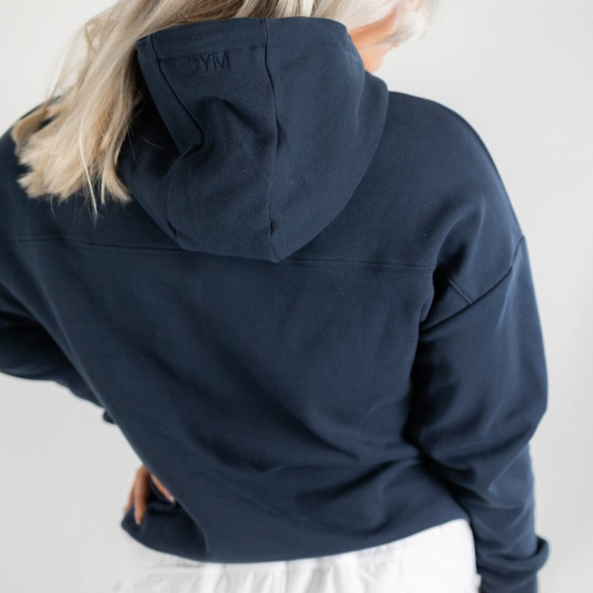A person with platinum blonde hair wears a DYM Athletics Cozy Up Cowl Neck Hoodie in ultra soft navy blue, GYM embroidered on the hood. Seen from the back with hand on hip, white garment peeking below, against a plain white background.