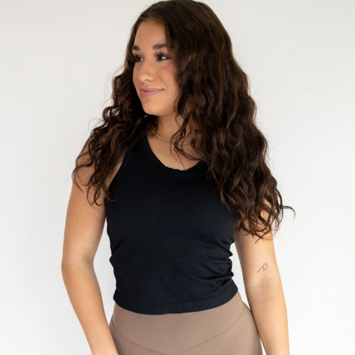 A woman with long, wavy brown hair stands against a white background wearing the DYM Athletics Restore Ribbed Comfort Tank w/ Built in Bra and high-waisted taupe leggings. She smiles softly, showing a small tattoo on her left forearm.