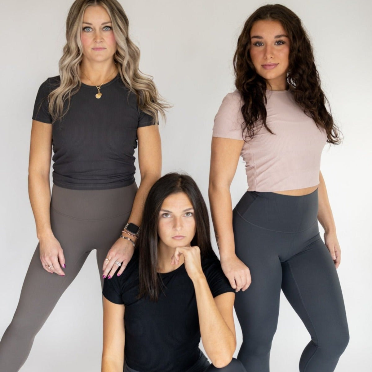 Three women pose confidently in DYM Athletics Restore Ribbed T-shirts and leggings. Two stand while one kneels, all wearing neutral tones with relaxed expressions, styled hair, and a strong modern look.