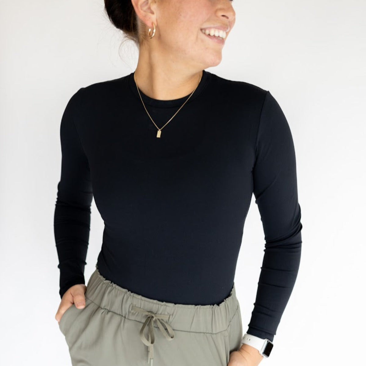 A person smiles with hands in pockets against a white background, wearing the DYM Athletics Lavish Shaping Bodysuit in black, olive drawstring pants, a gold necklace, small hoop earrings, and a smartwatch. Only the upper body is shown.