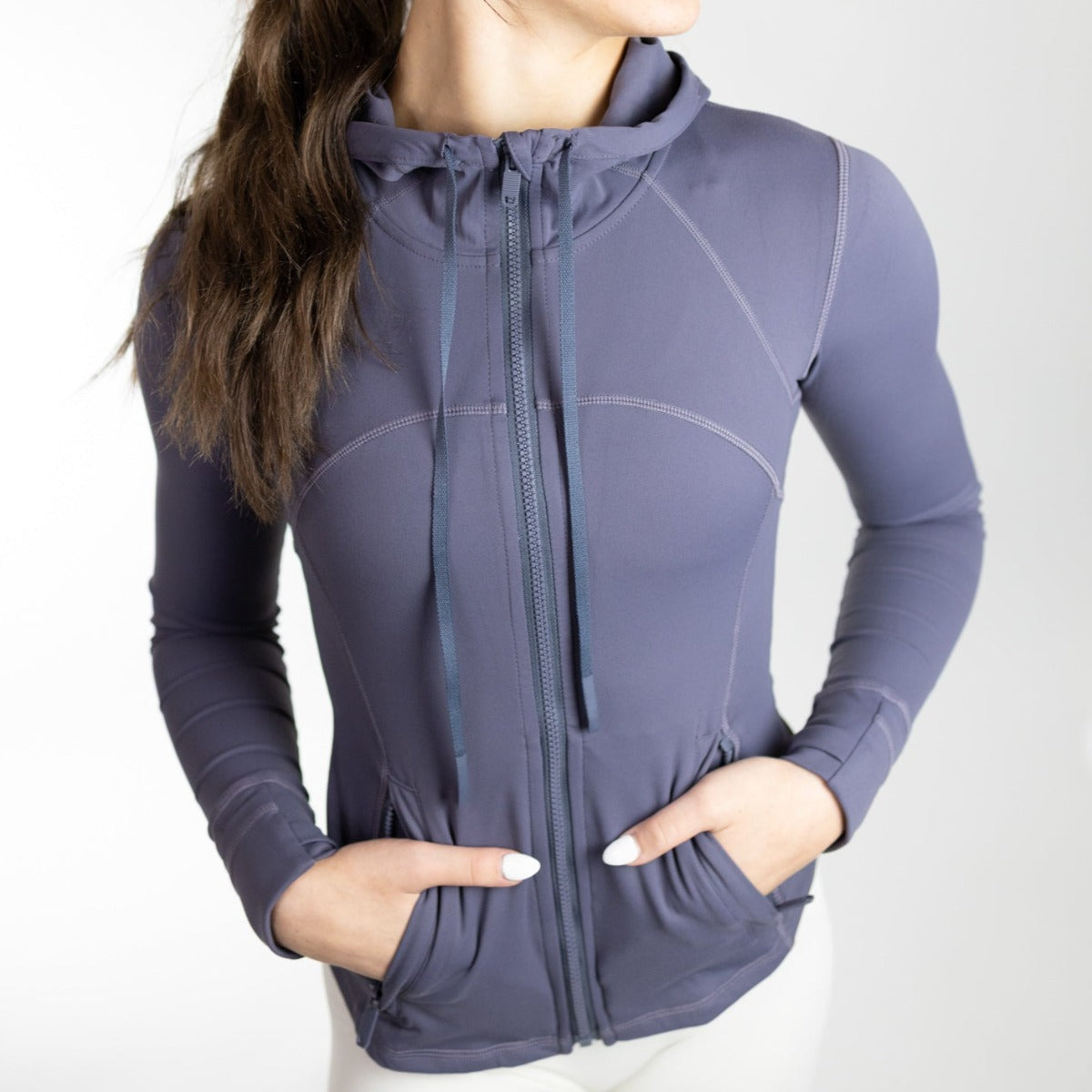 A woman stands with hands in pockets, wearing the DYM Athletics Active Lightweight Hooded Zip Up in slate blue. This fitted, long-sleeve jacket features four-way stretch and visible seams—perfect for an active lifestyle. She faces slightly away from the camera.