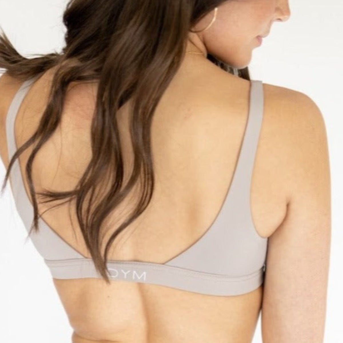 A woman with long brown hair is seen from the back wearing a light beige Luxe Lift Everyday Bra by DYM Athletics, featuring thin straps and “DYM” on the band, offering wire-free comfort and light support against a plain background.