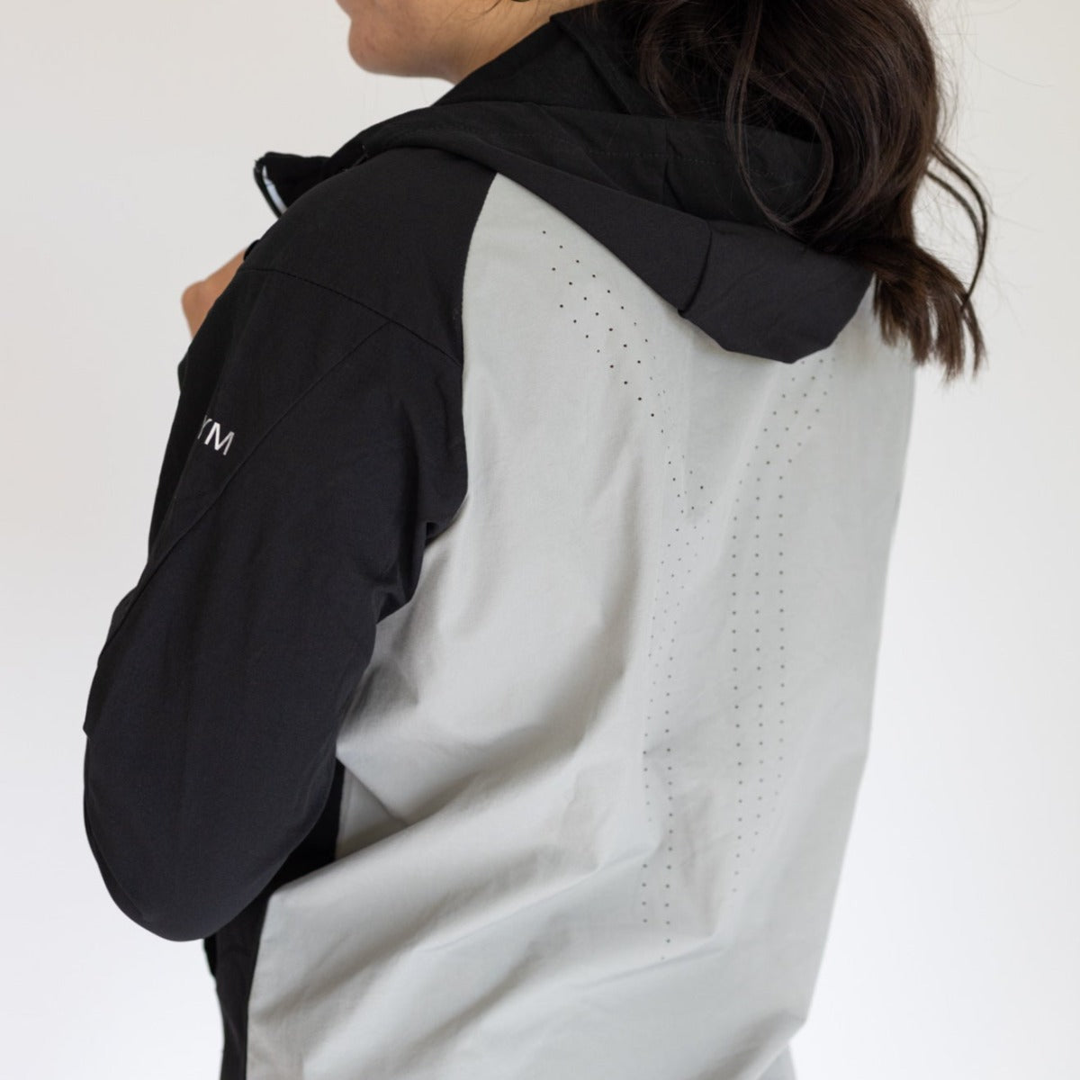 A woman with long dark hair wears the DYM Athletics Women's Hiking Jacket in light gray and black, featuring upper back ventilation holes and a sleeve logo. The water resistant jacket is shown against a plain white background.