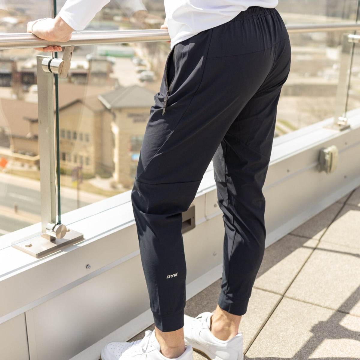 A person stands on a sunlit rooftop balcony wearing DYM Athletics' Fly Lightweight Active Jogger | SALE, paired with a white long-sleeve shirt and white sneakers. One hand rests on the glass railing, with city buildings blurred in the background.