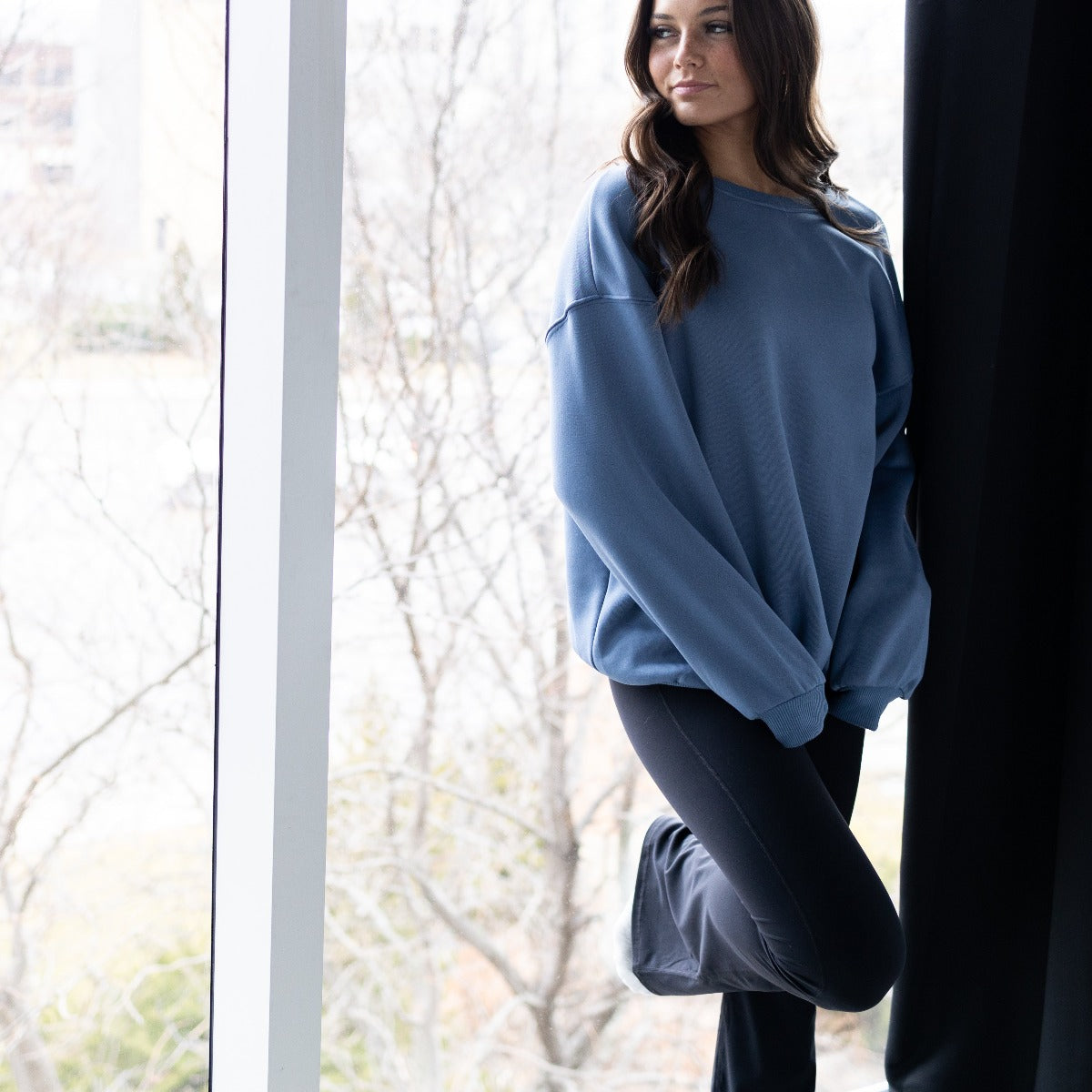 A woman in a loose blue sweatshirt and DYM Athletics AIRE Ultra-Soft Fabulous Flare High Waist Pant stands indoors by a large window, her activewear perfect for relaxing moments. Bare tree branches are visible outside.