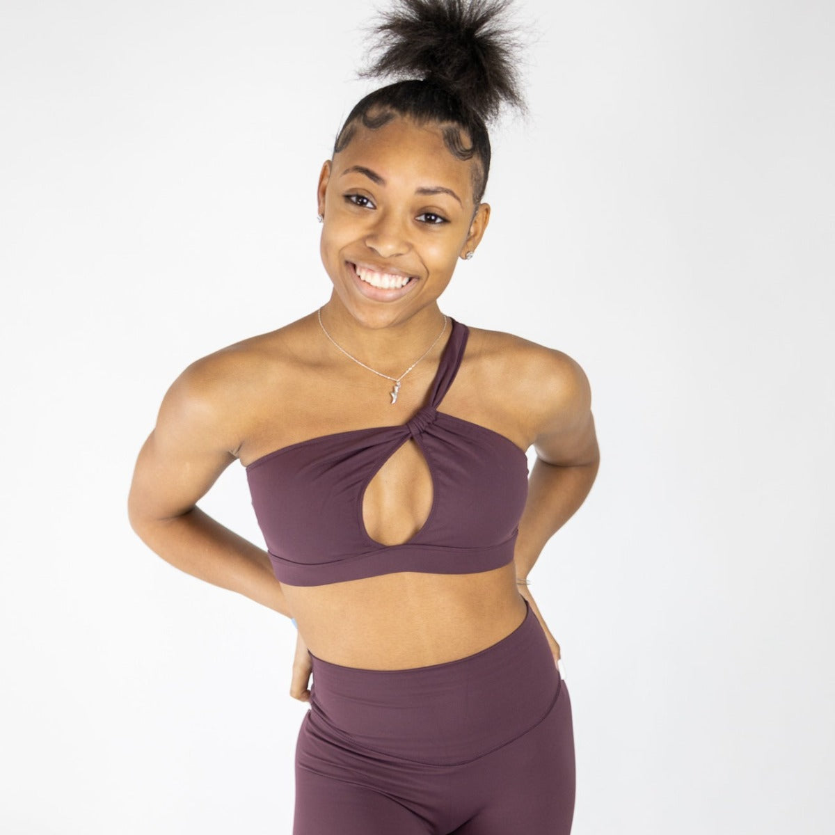 A young woman with a high bun smiles at the camera, wearing DYM Athletics' The Siren One Shoulder Cutout Bra (A-C Cup) and Muse Leggings. Ideal for barre or everyday wear, she poses against a plain white background with her hands behind her back.
