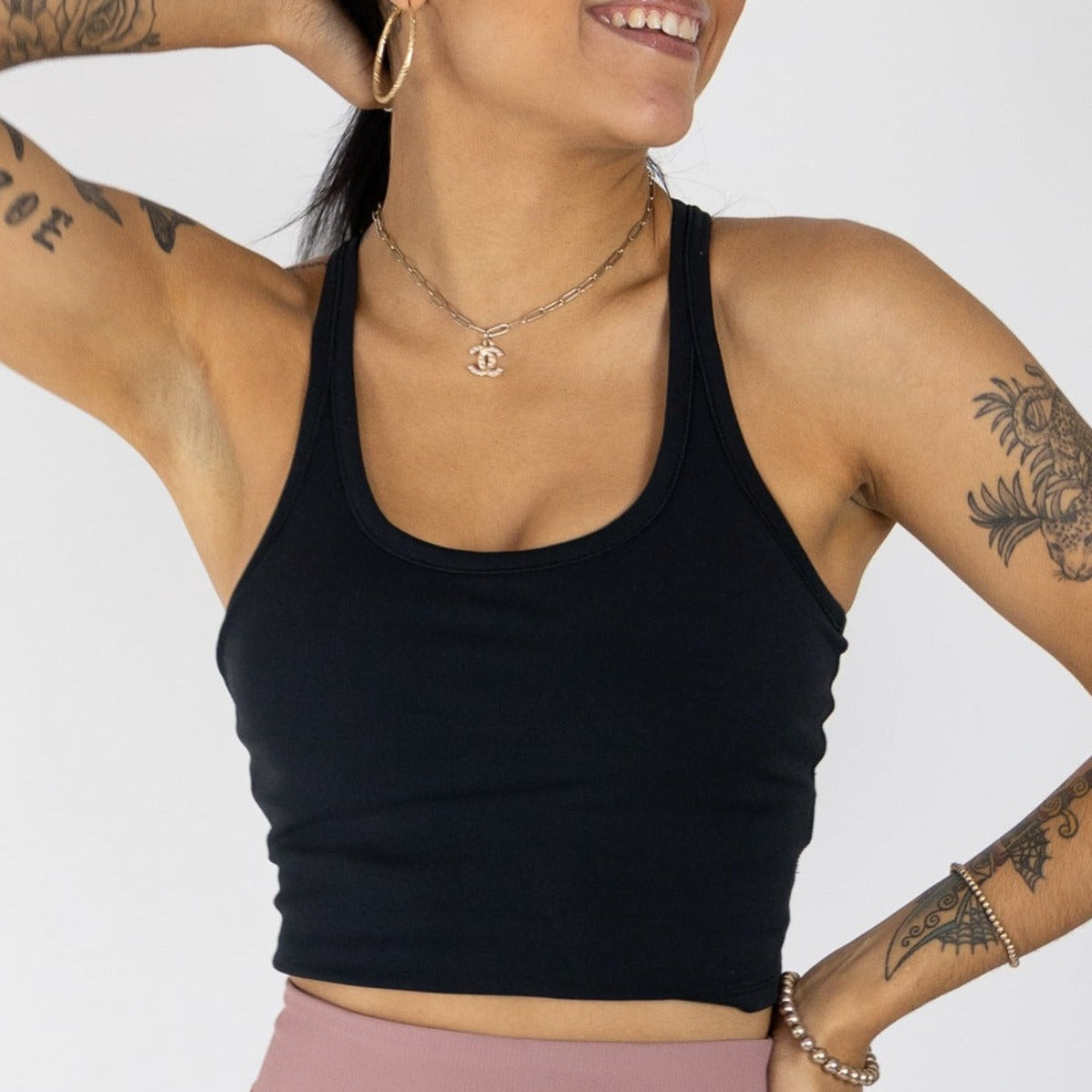 A smiling person wears the Energy Crop Everyday Tank by DYM Athletics, featuring a racerback and seamless design, paired with high-waisted mauve leggings. They accessorize with gold hoops, a pendant necklace, and pose against a light gray background.