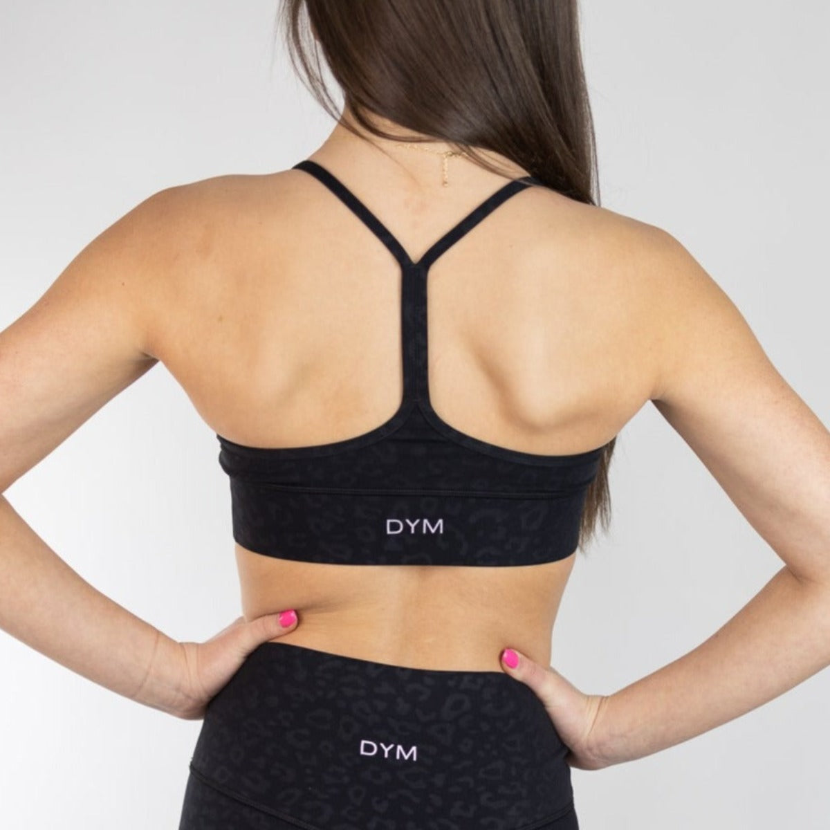 A woman with her back to the camera wears the DYM Athletics Flow Bra (A-C cup, medium support) and matching black high-waisted leggings. Her brown hair is down and she has pink nail polish. The background is plain and light.