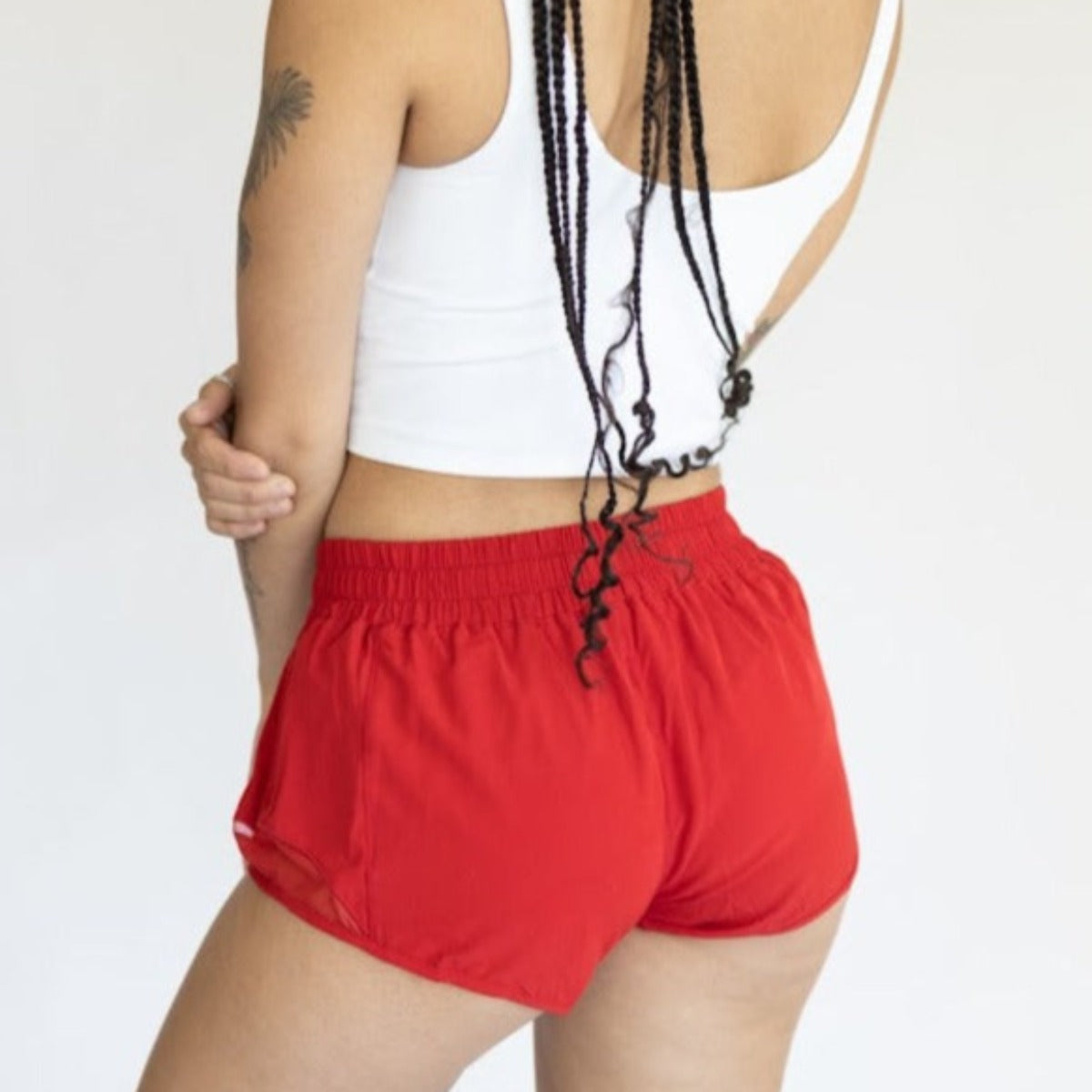 A person with long braids wears a white sleeveless top and red DYM Athletics Chic Athletic Short | 1.5" with an adjustable drawstring, facing away from the camera against a plain light background, one hand on their hip.