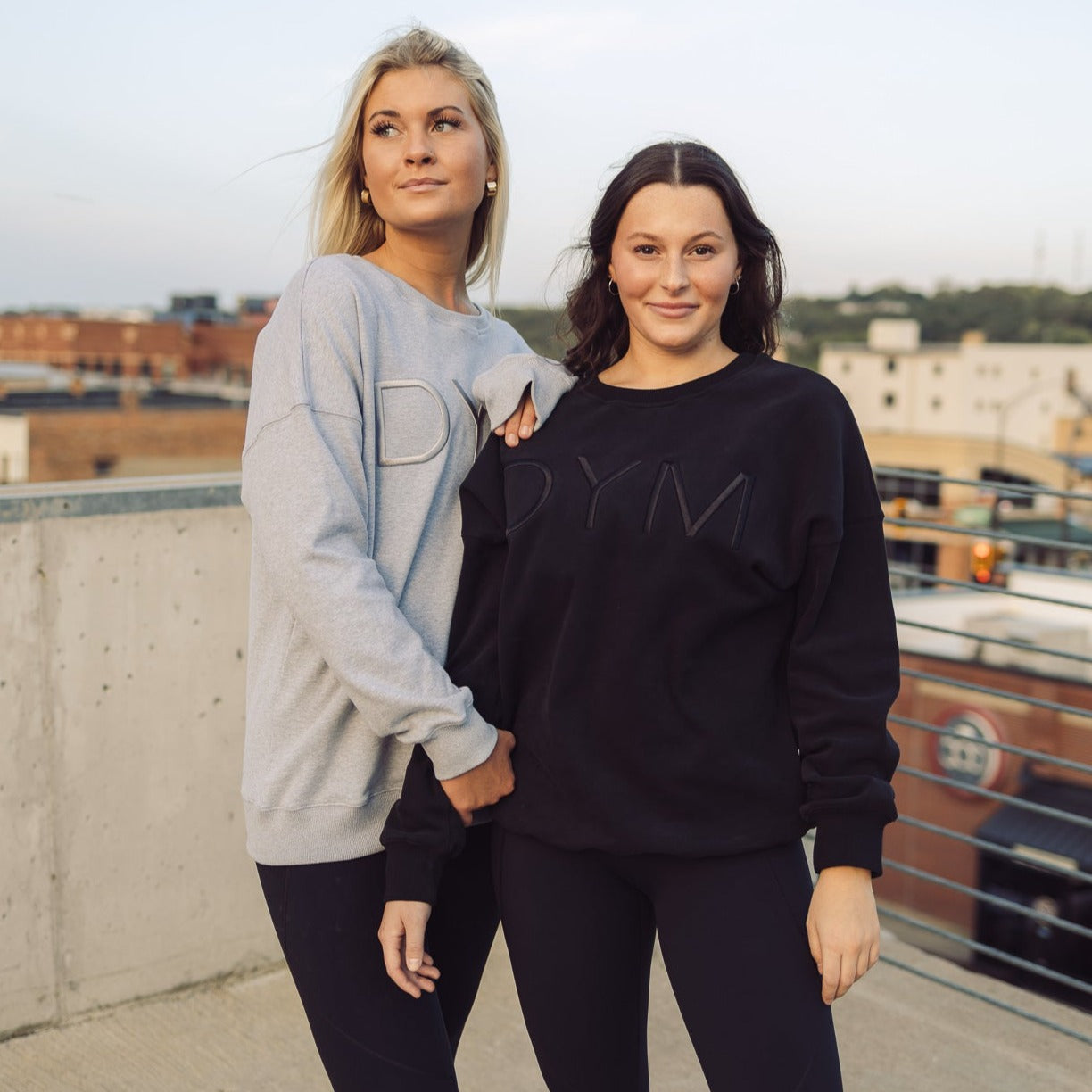 Two women stand on a rooftop in DYM Athletics’ Cozy Up Crew Neck Sweatshirts. Their confident smiles and the ultra-soft, relaxed fit show comfort, as sunset clouds and city buildings frame them in the background.