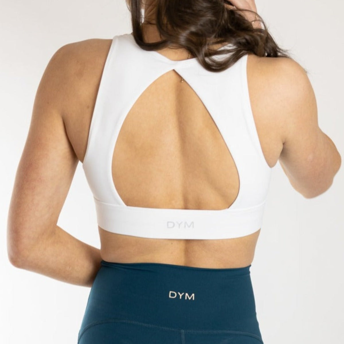 A woman is seen from behind in the DYM Athletics Heartbreaker Deep V Sports Bra with Keyhole Back, featuring a white design and “DYM” on the band. She pairs it with high-waisted teal DYM leggings for a chic activewear look, her long hair draped over one shoulder.