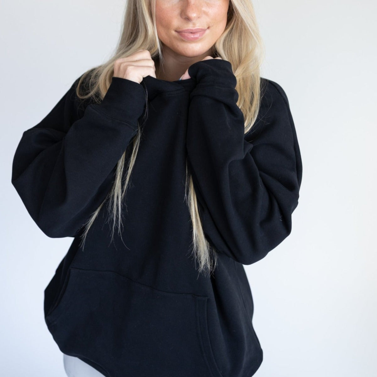 A person with long blonde hair wears the oversized black Cozy Up Boyfriend Hoodie by DYM Athletics, holding the collar with both hands. They stand against a light background, upper body visible, face partly hidden by the cozy fabric.