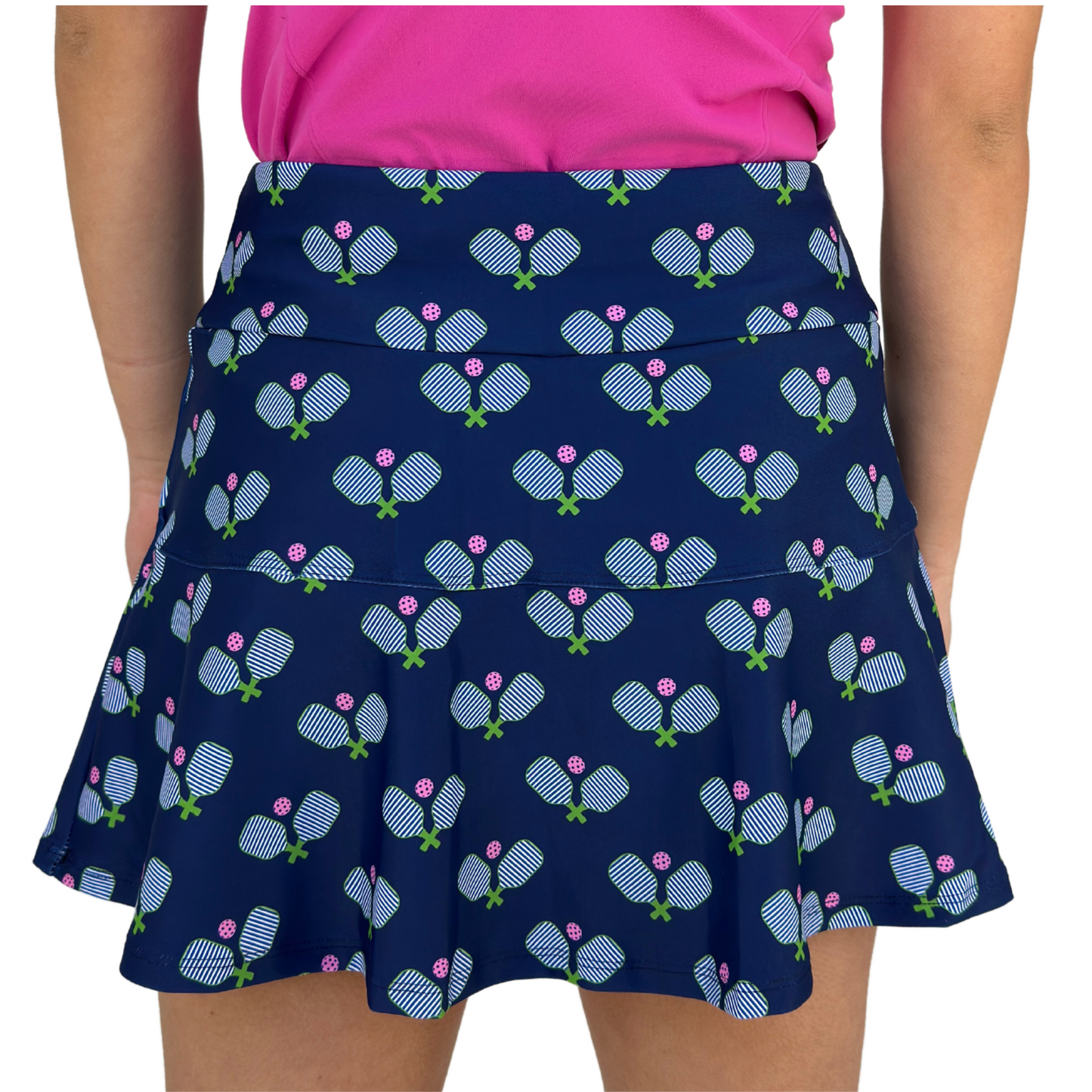 A person wears the Hit It! Pickleball Skirt by Queen of the Court, featuring a flared hem and a print of white and blue paddles with green handles and pink balls. It's paired with a bright pink shirt, all proudly made in the USA.