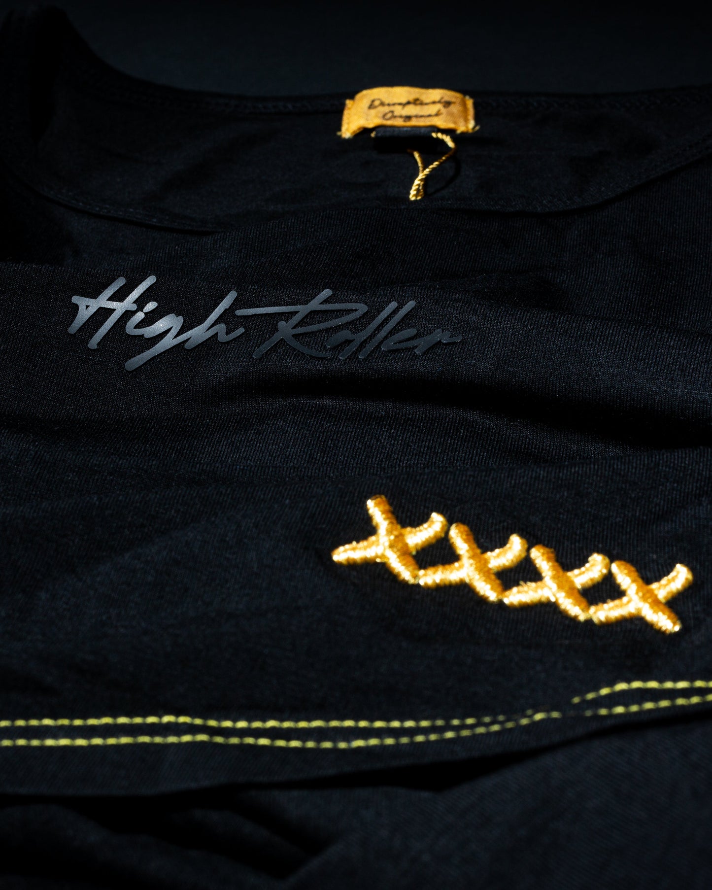 A close-up of the black High Roller Lounge Pickleball T-Shirt by Dope Pickleball Co. shows silver cursive text, yellow X stitching, bold yellow seams, textured fabric, and a tan neck label—ideal for fans of the brand's signature style.