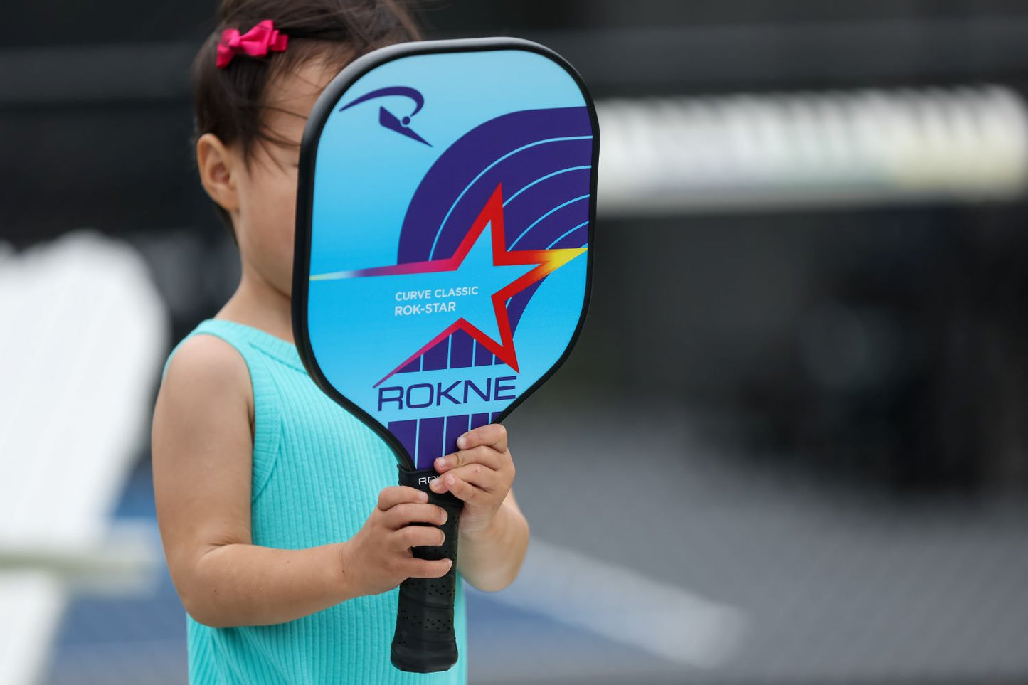 A young child in a turquoise sleeveless outfit holds a ROKNE CURVE CLASSIC ROK-STAR (KIDS PADDLE) featuring a blue, purple, and red star design, obscuring most of their face on an outdoor pickleball court.