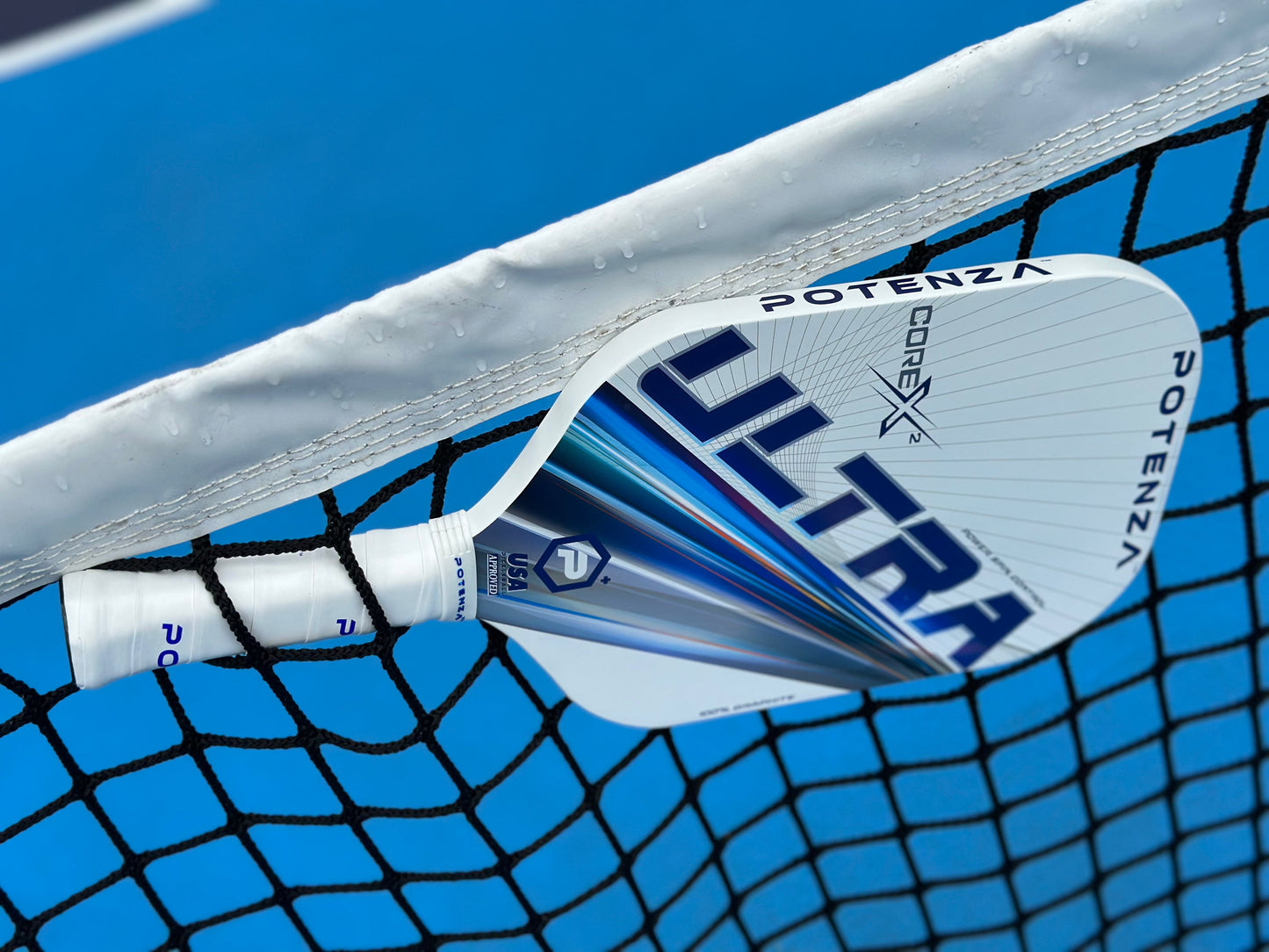 A close-up of the Potenza Pickleball ULTRA+ COREx2 edgeless paddle in white and blue rests on a black net over a blue court. Water droplets sparkle on the paddle and net, with clear court lines visible in the background.