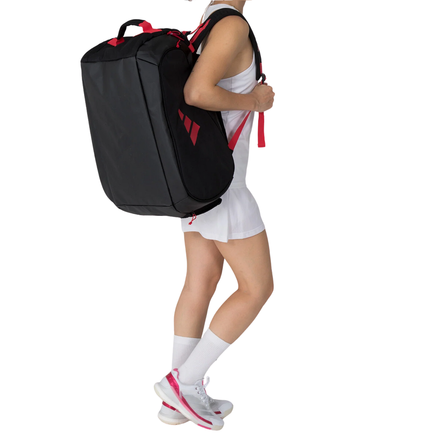 A person in a white tennis outfit and pink-accented sneakers carries the Adidas TOUR BLACK Racket Bag 3.4 by Padel USA, featuring red straps, over one shoulder. The person's head is not visible against a plain white background.