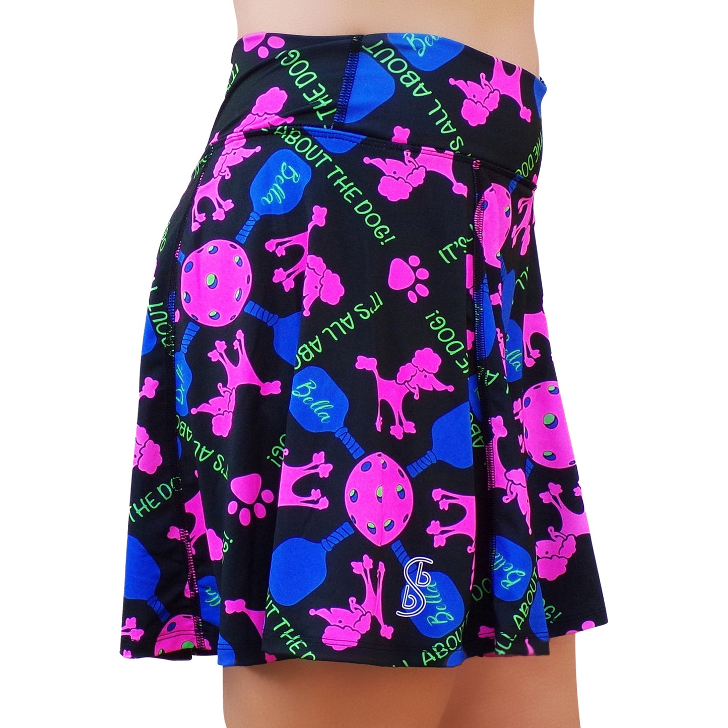 A woman wears the Pickleball Bella PB Aline Skort Pickles, a vibrant athletic skort with neon pink dog silhouettes, paw prints, blue bones, green tennis balls on black, and “ITS ALL ABOUT THE DOG!” text in green.