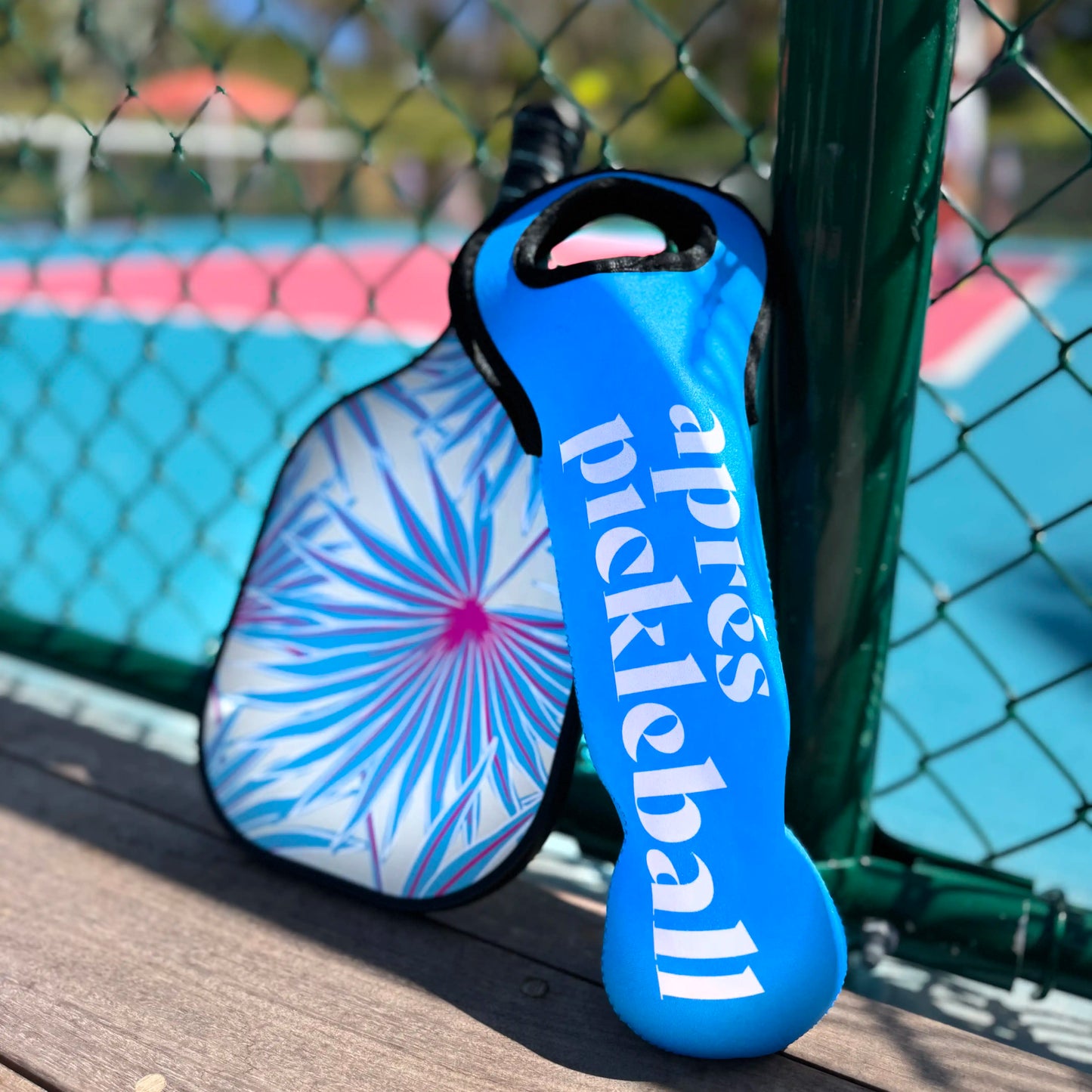 Two Swinton Pickleball paddles rest by a court and green fence, with an Aprés Pickleball Sleeve of Pickleballs bag nearby. One paddle’s cover says “après pickleball,” the other features a vibrant blue, pink, and white starburst pattern.