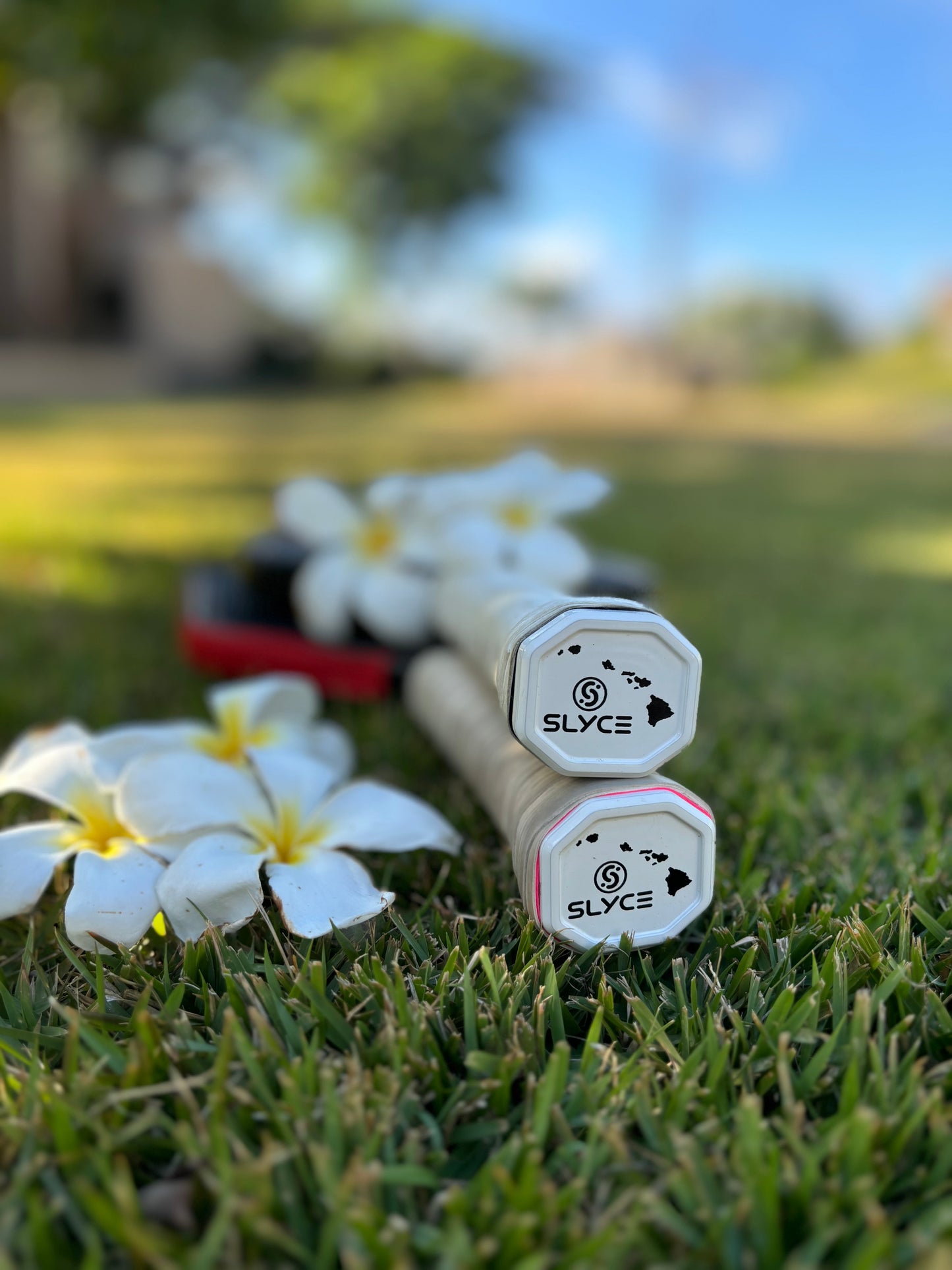 Two Slyce Sport Special Edition Hawaii Islands SpeedCap (28g, 1.0 oz.) Paddle Performance Enhancer products with white handles rest on green grass beside white plumeria flowers, while a red paddle case sits in the sunny outdoor background.