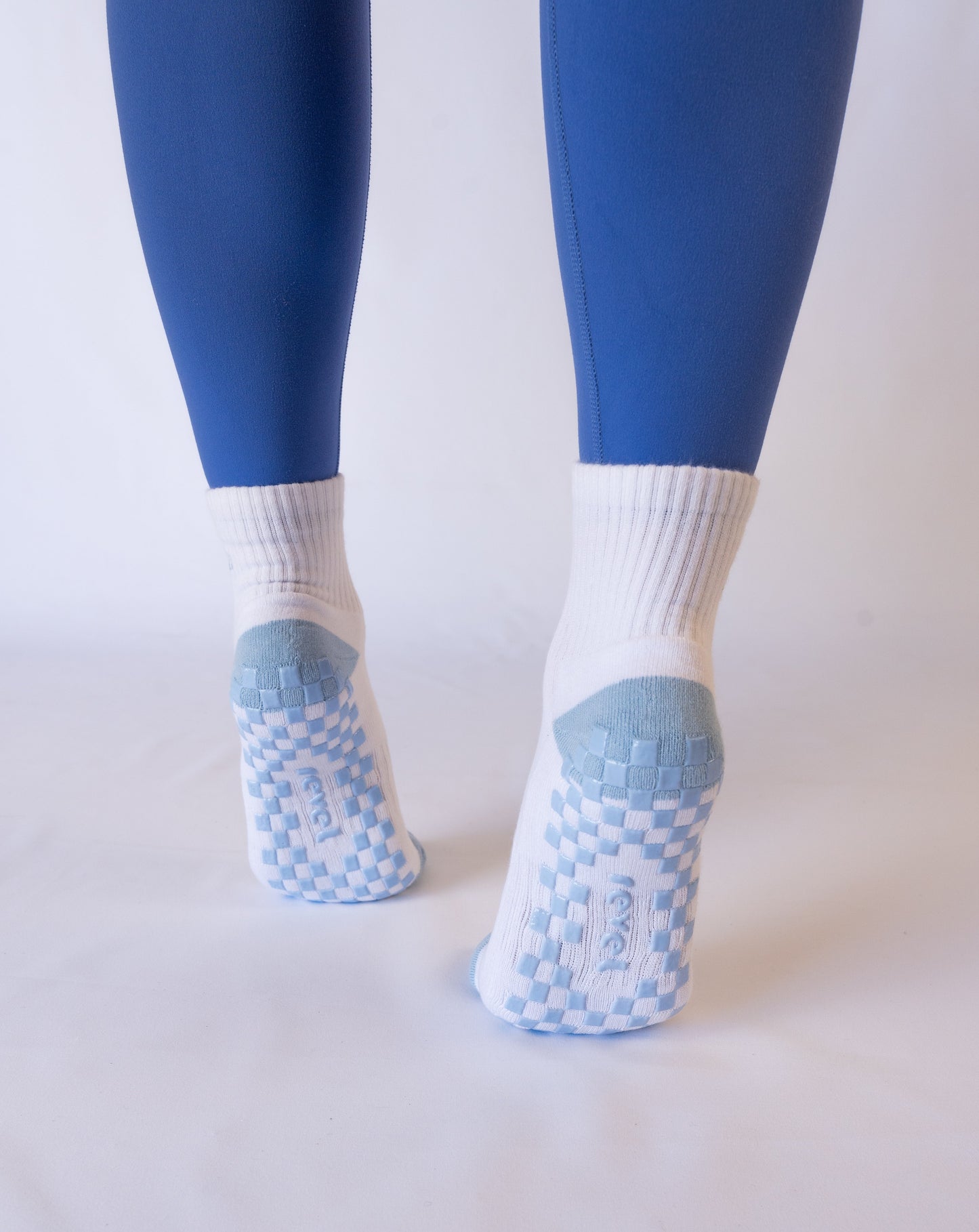 A person wears Revel’s The Pardon My French Grip Sock—white ankle socks with blue and white checkered non-slip soles—paired with blue leggings, standing on a white surface with heels lifted and a plain light background highlighting their feet.