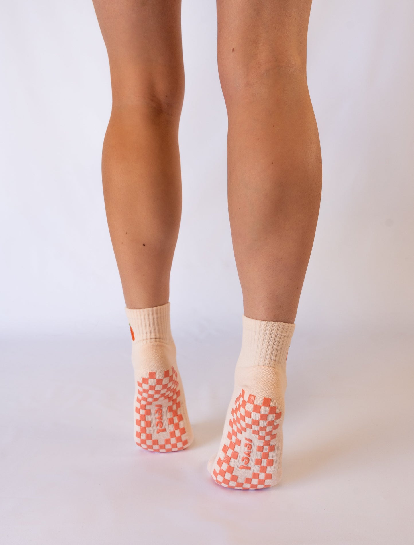 A person stands on tiptoe, displaying the backs of their legs in Revel’s The Just Peachy Grip Sock. These cream Pilates socks feature orange checkered non-slip soles with “FRESH” text, shown against a plain white background for focus.