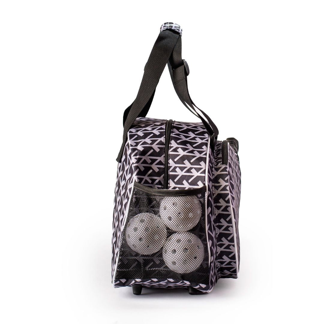 The Court Couture Black Premium Women's Pickleball Bag by Pik'le'Ball Sports is displayed upright, featuring a black-and-white geometric pattern, zipper top, handle, long black strap, and three white pickleballs in a mesh side pocket.