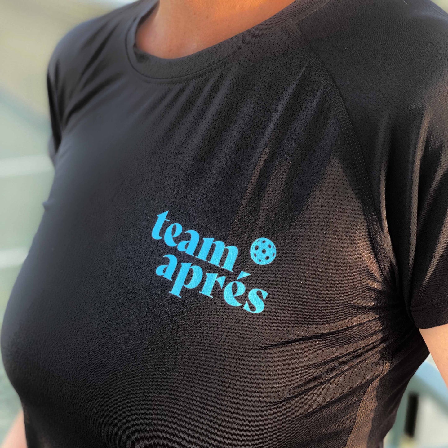 Close-up of Swinton Pickleball’s Women's Aprés Pickleball Performance Shirt—black, with “team après” in light blue and a pickleball graphic above the "e." Moisture-wicking fabric keeps you comfortable on the court. Outdoor sports setting in background.