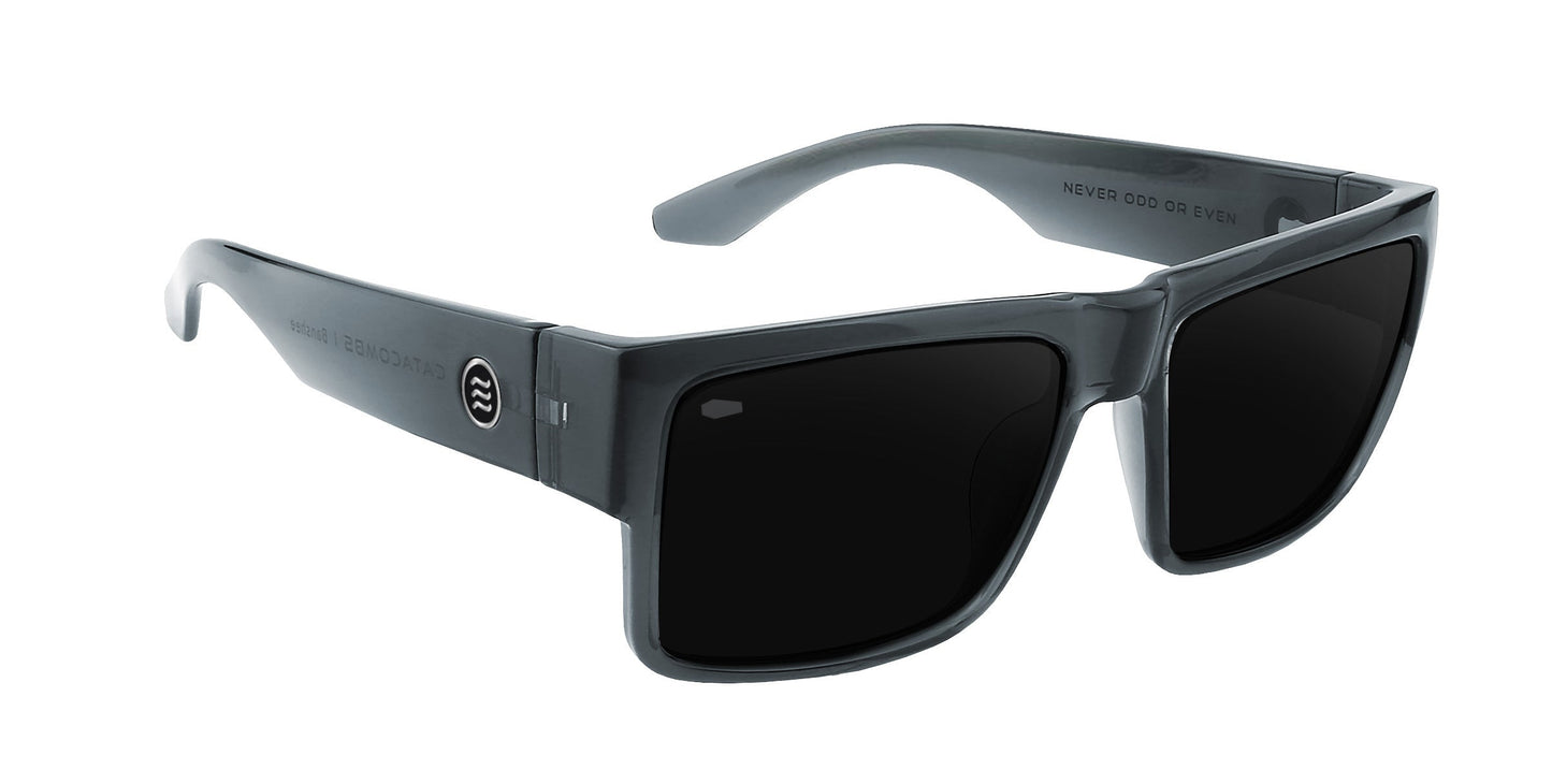 The Banshee by Neven Eyewear features black rectangular frames with dark lenses, a round logo on the left arm, and “NEVER ODD OR EVEN” printed inside the right—modern, sporty sunglasses from the Catacombs collection.