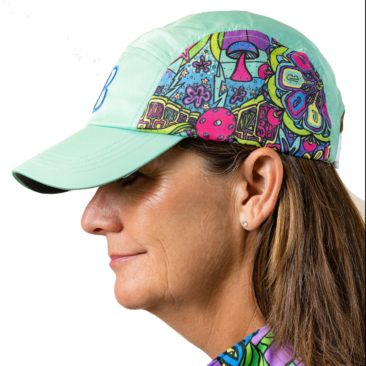 A woman in profile wears the Pickleball Bella PB Cap Groovy, a mint-green baseball hat with colorful psychedelic mushrooms and floral designs. She has light brown hair, stud earrings, and a patterned shirt against a plain white background.