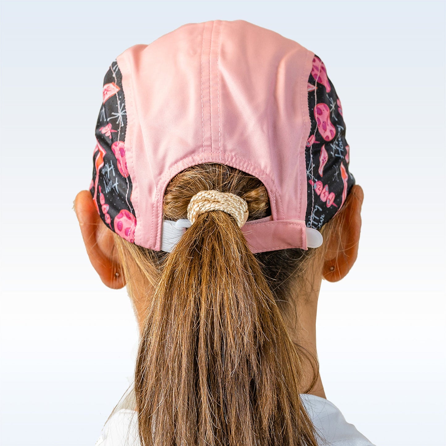 A woman seen from behind wears a Pickleball Bella PB Cap Martini 2-4-1 Happy Hour scrub cap with pink skull and bone patterns, her light brown hair pulled through the adjustable velcro ponytail hole. She wears a white-collared top.