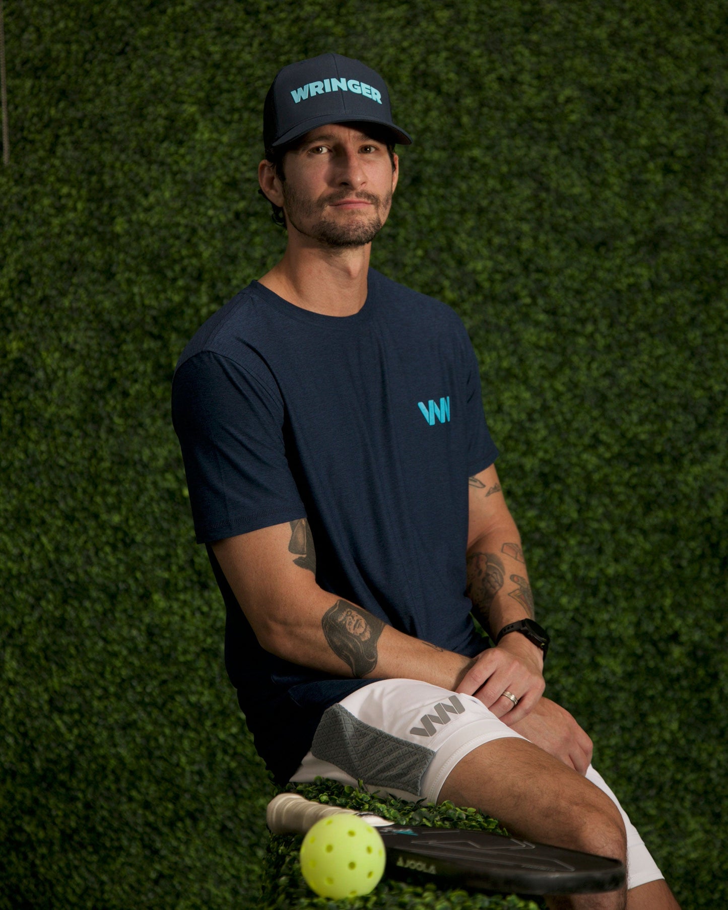 A tattooed man in white shorts wears a navy Adjustable Mesh WRINGER Hat by Wringer Wear, featuring a snapback closure, as he sits against green foliage beside a paddle and yellow perforated ball, suggesting paddle sports.