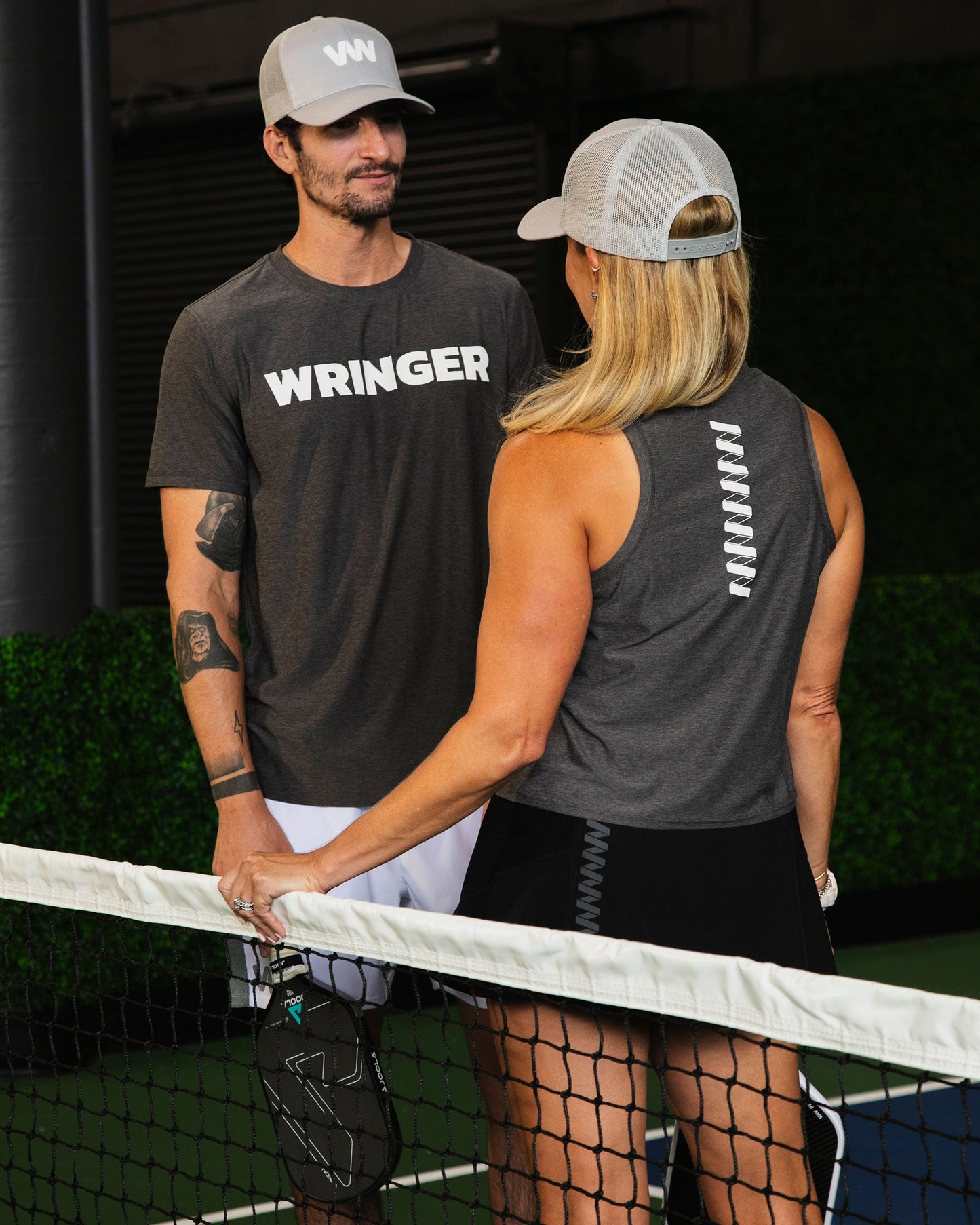 A man and woman stand on opposite sides of a tennis net. The woman, in a Wringer Wear Women's Performance Logo Tank, holds a pickleball paddle. Both wear slim sportswear and gray hats on an indoor court with lush greenery.