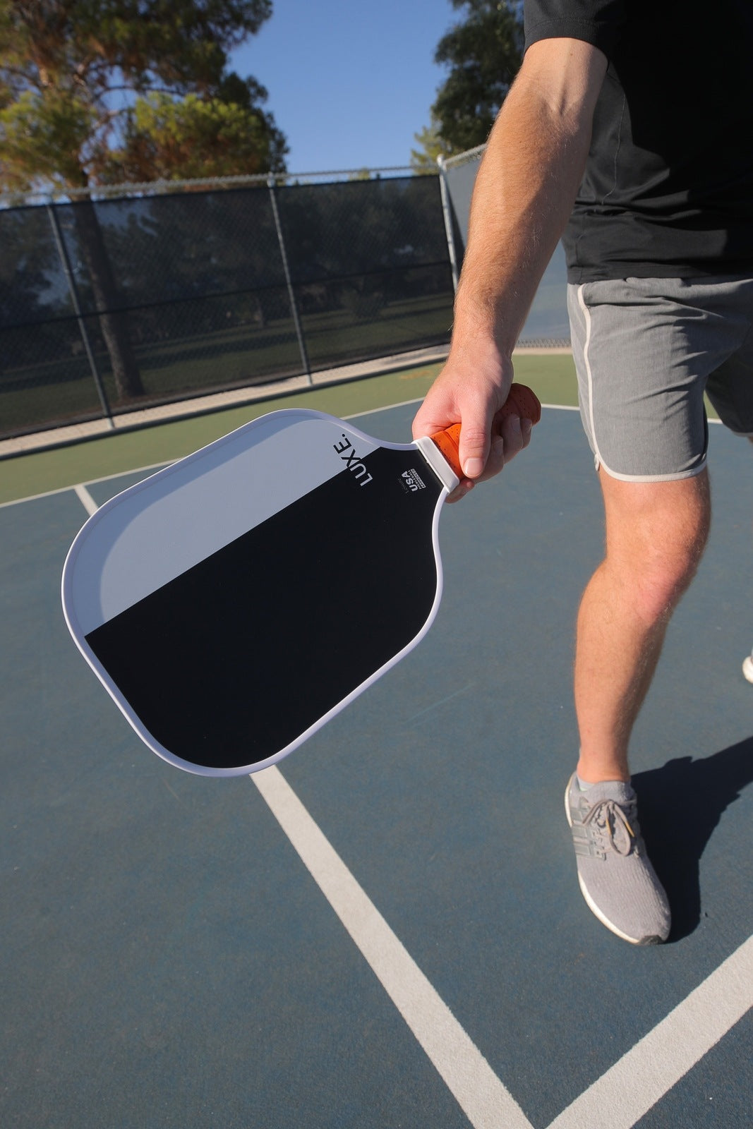 A person in a black shirt and gray shorts holds the LUXE Pickleball Dipped – Leisure paddle with a fiberglass surface over a blue outdoor court, with only their lower body and arm visible against the fence, trees, and sky.