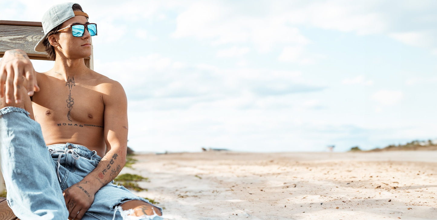 A shirtless person in ripped jeans and a backward cap relaxes on a beach bench, wearing Neven Eyewear’s Melby polarized sunglasses. Tattoos are visible on their chest and arms. The sky is bright and cloudy, with sand stretching out behind them.